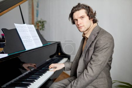 Téléchargez les photos : Portrait de pianiste professionnel en costume regardant la caméra tout en jouant des notes sur le piano à l'école de musique - en image libre de droit