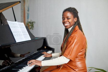 Photo for Portrait of African young woman smiling at camera while sitting at piano, she happy to learn playing the piano - Royalty Free Image