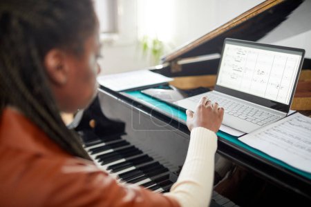 Foto de Vista trasera de la mujer africana mirando partituras en el monitor de su computadora portátil mientras aprende a tocar el piano - Imagen libre de derechos