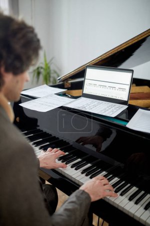 Téléchargez les photos : Vue arrière du jeune homme jouant du piano à l'aide d'un ordinateur portable avec une partition sur l'écran pendant la leçon en ligne - en image libre de droit