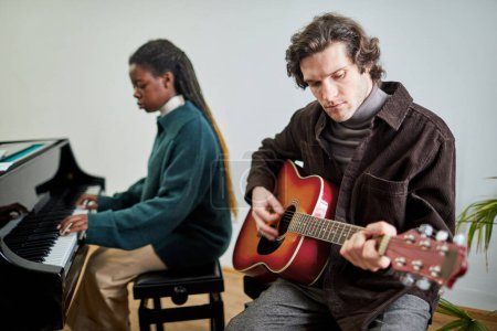Foto de Joven sentado en la silla y tocando la guitarra con la mujer africana tocando el piano en el fondo, que tocando instrumentos musicales en dúo - Imagen libre de derechos
