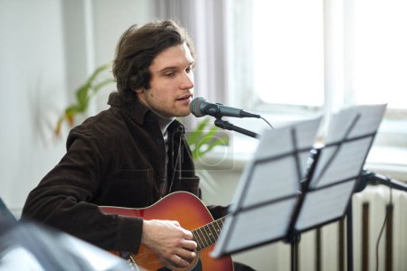 Téléchargez les photos : Jeune homme regardant la partition et chantant une chanson au microphone tout en jouant de la guitare en studio - en image libre de droit