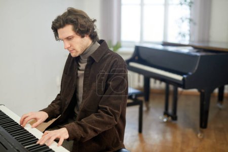 Téléchargez les photos : Jeune homme jouant du piano et apprenant la nouvelle mélodie assis en classe à l'école de musique - en image libre de droit