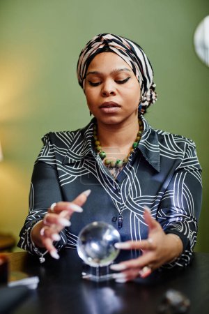 Photo for Vertical portrait of ethnic fortune teller gazing into crystal ball at spiritual seance in shop - Royalty Free Image