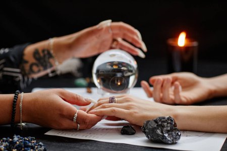 Téléchargez les photos : Gros plan de diseur de bonne aventure tenant la main d'une jeune femme lors d'une séance spirituelle avec une boule de cristal - en image libre de droit