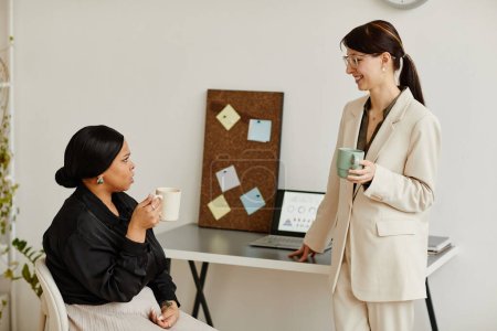 Téléchargez les photos : Portrait minimal de deux jeunes femmes qui ont réussi à parler à la pause café dans leur bureau et à tenir des tasses - en image libre de droit