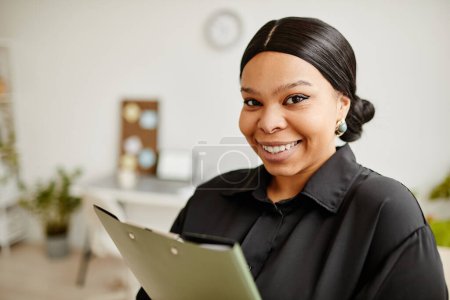 Téléchargez les photos : Gros plan portrait de succès femme d'affaires noire souriant à la caméra tout en se tenant dans un bureau minimal - en image libre de droit