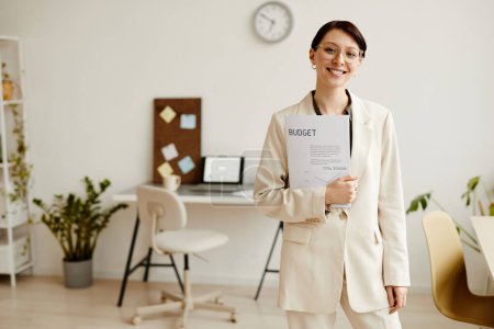 Foto de Retrato de cintura hacia arriba de la exitosa joven empresaria sonriendo a la cámara mientras está de pie en una oficina mínima, espacio para copiar - Imagen libre de derechos