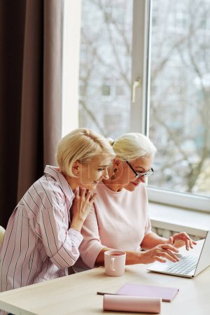 Téléchargez les photos : Portrait latéral de jeune femme avec mère aînée utilisant l'ordinateur ensemble contre la fenêtre à la maison - en image libre de droit