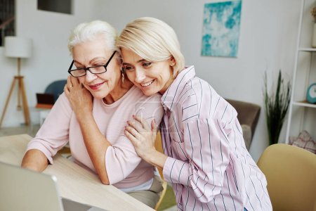 Foto de Retrato de mujer joven sonriente abrazando a la madre mayor mientras miran juntos a la pantalla del portátil en el acogedor interior del hogar - Imagen libre de derechos