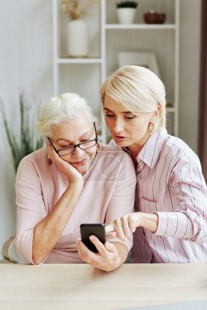 Téléchargez les photos : Portrait vertical de jeune femme blonde aidant la mère aînée à utiliser un smartphone dans un intérieur confortable - en image libre de droit