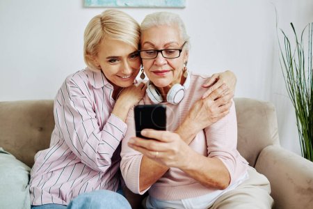 Téléchargez les photos : Portrait de femme âgée moderne utilisant un appareil smartphone à la maison avec une jeune fille souriante l'embrassant - en image libre de droit