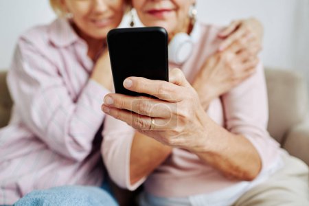 Téléchargez les photos : Gros plan d'une femme âgée méconnaissable tenant un smartphone avec sa fille l'embrassant - en image libre de droit