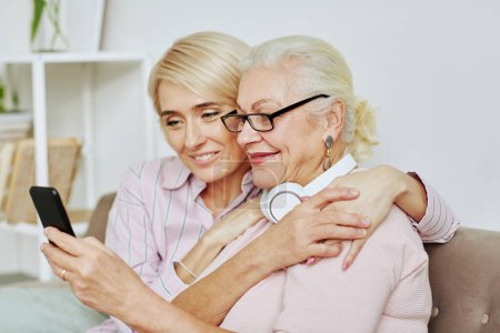 Téléchargez les photos : Portrait de fille adulte souriante embrassant la mère aînée et regardant l'écran du smartphone dans la maison confortable - en image libre de droit