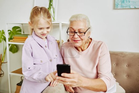 Téléchargez les photos : Portrait de mignonne petite fille avec grand-mère regardant l'écran du smartphone dans la maison confortable - en image libre de droit
