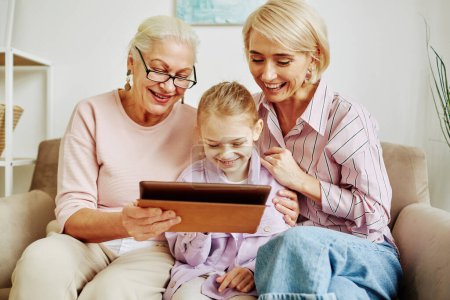 Téléchargez les photos : Portrait de la famille heureuse de toutes les femmes regardant l'écran de la tablette numérique et souriant tout en étant assis sur le canapé ensemble - en image libre de droit
