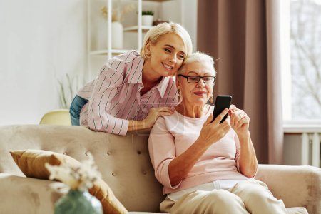 Photo for Portrait of smiling adult daughter with mother using smartphone together in cozy home scene - Royalty Free Image