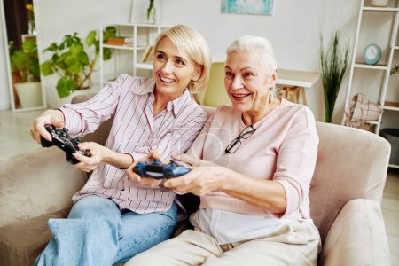 Foto de Retrato de una mujer mayor sonriente jugando videojuegos con una hija adulta en una acogedora escena en casa - Imagen libre de derechos