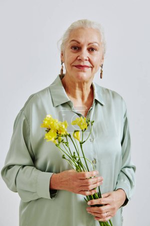 Téléchargez les photos : Portrait taille haute minime de femme âgée aux cheveux blancs tenant des fleurs et regardant la caméra en studio - en image libre de droit