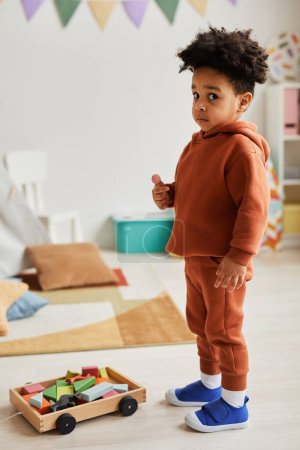 Téléchargez les photos : Portrait vertical pleine longueur de mignon bambin afro-américain regardant la caméra tout en jouant avec des jouets dans la chambre des enfants - en image libre de droit