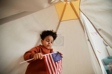 Foto de Retrato de ángulo bajo del niño negro lindo que sostiene la bandera estadounidense mientras juega en la tienda de campaña de los niños, espacio de copia - Imagen libre de derechos