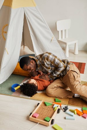Foto de Retrato vertical del amoroso padre negro jugando con su pequeño hijo en el suelo en la acogedora habitación de los niños, ambos riendo felizmente - Imagen libre de derechos