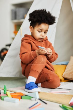 Téléchargez les photos : Portrait vertical pleine longueur d'un jeune garçon afro-américain jouant sur le sol dans une chambre d'enfant habillé en tenue neutre à la mode - en image libre de droit
