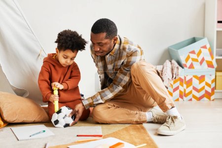 Photo for Full length portrait of caring black father playing with toddler son indoors and pumping up kids soccer ball, copy space - Royalty Free Image