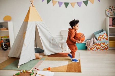 Foto de Retrato de cuerpo entero de un lindo niño afroamericano sosteniendo una pelota de baloncesto mientras juega en casa en una acogedora habitación para niños y vistiendo un atuendo de moda mínimo - Imagen libre de derechos