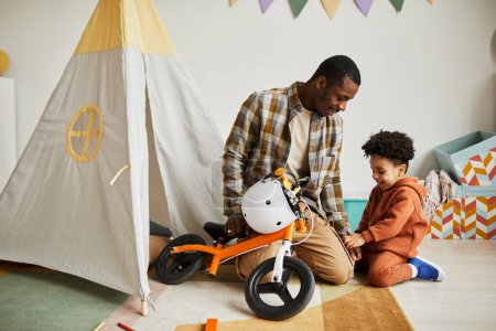 Foto de Retrato de cuerpo entero del joven padre negro jugando con su hijo pequeño en el interior y enseñándole a montar en primera bicicleta de equilibrio - Imagen libre de derechos