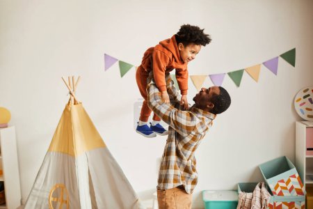 Téléchargez les photos : Vue latérale minime portrait d'un père noir aimant jouant avec un tout-petit fils à la maison et le tenant dans les airs, espace de copie - en image libre de droit