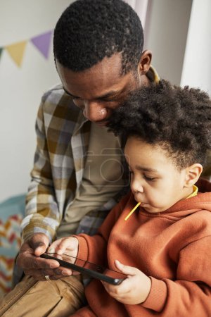 Foto de Retrato vertical del padre negro y el pequeño hijo lindo usando el teléfono inteligente juntos y viendo dibujos animados en línea - Imagen libre de derechos
