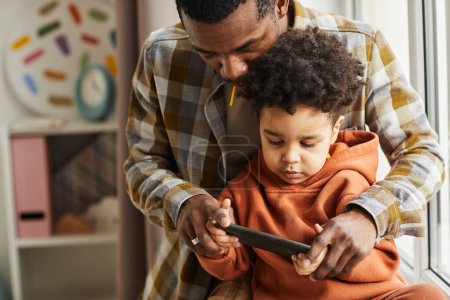 Foto de Retrato de la cintura hacia arriba del padre y el niño afroamericanos usando un teléfono inteligente juntos y viendo dibujos animados en línea, espacio para copiar - Imagen libre de derechos
