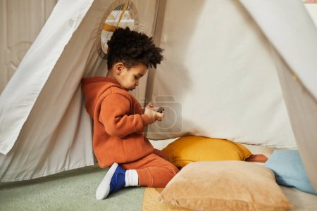 Photo for Minimal side view portrait of little African American boy using smartphone in play tent and wearing trendy outfit, gen Alpha, copy space - Royalty Free Image