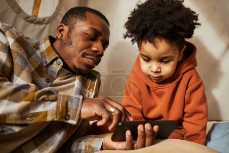 Téléchargez les photos : Portrait de père noir avec un petit garçon en bas âge utilisant un smartphone ensemble et regardant des dessins animés dans une tente de jeu - en image libre de droit