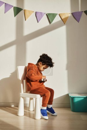 Photo for Minimal full length portrait of little toddler using smartphone while sitting on chair in kids room lit by sunlight - Royalty Free Image