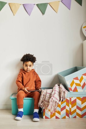 Foto de Retrato mínimo de longitud completa de niño negro sosteniendo la tableta y mirando a la cámara en el interior de la habitación de los niños lindo - Imagen libre de derechos