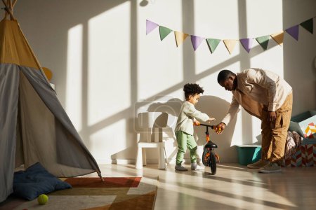 Photo for Minimal portrait of loving black father teaching little boy riding balance bike at home in sunlight, copy space - Royalty Free Image