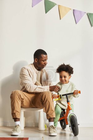 Téléchargez les photos : Minimal portrait pleine longueur de jeune père noir enseignant petit garçon équitation équilibre vélo à la maison - en image libre de droit