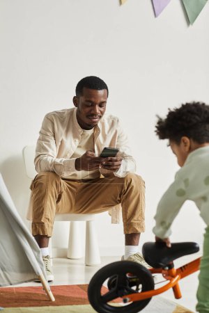 Photo for Vertical portrait of young black father using smartphone while watching little boy at home - Royalty Free Image