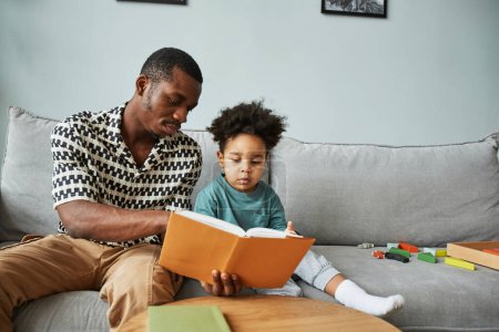 Foto de Retrato del padre negro y el hijo leyendo libros juntos mientras están sentados en el sofá en casa, espacio para copiar - Imagen libre de derechos