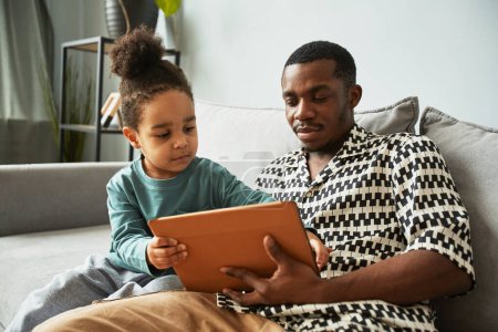 Foto de Retrato de un joven padre negro y un niño pequeño usando una tableta digital juntos mientras se relajan en el sofá en casa - Imagen libre de derechos