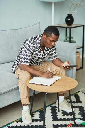Photo for Full length portrait of young black man working from home and using smartphone in grey interior - Royalty Free Image