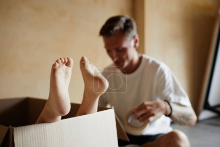 Foto de Primer plano de lindos pies de bebé que sobresalen de la caja de cartón mientras la familia se muda de casa en el fondo, espacio para copiar - Imagen libre de derechos