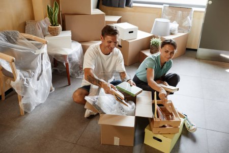Photo for Portrait of tattooed couple unpacking cardboard boxes sitting on floor while moving into new house, copy space - Royalty Free Image