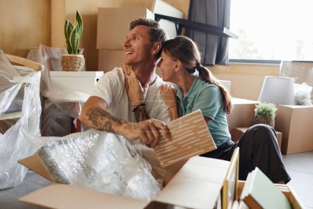Photo for Portrait of happy tattooed couple unpacking boxes sitting on floor while moving into new house, copy space - Royalty Free Image