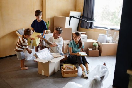 Photo for Full length portrait of happy family with two children unpacking boxes together while moving house, copy space - Royalty Free Image
