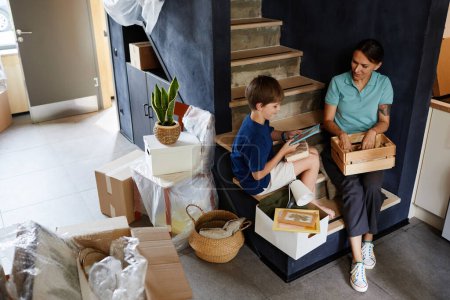 Foto de Retrato de larga duración de la madre y el hijo desempacando cajas mientras están sentados en escaleras en un nuevo hogar y sonriendo, espacio para copiar - Imagen libre de derechos
