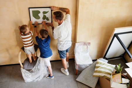 Foto de Retrato de vista superior del padre con dos hijos colgando una foto en la pared mientras se mudan a un nuevo hogar, espacio para copiar - Imagen libre de derechos