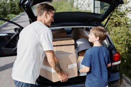 Photo for Back view portrait of father and son loading boxes to car while moving into new house lit by sunlight - Royalty Free Image